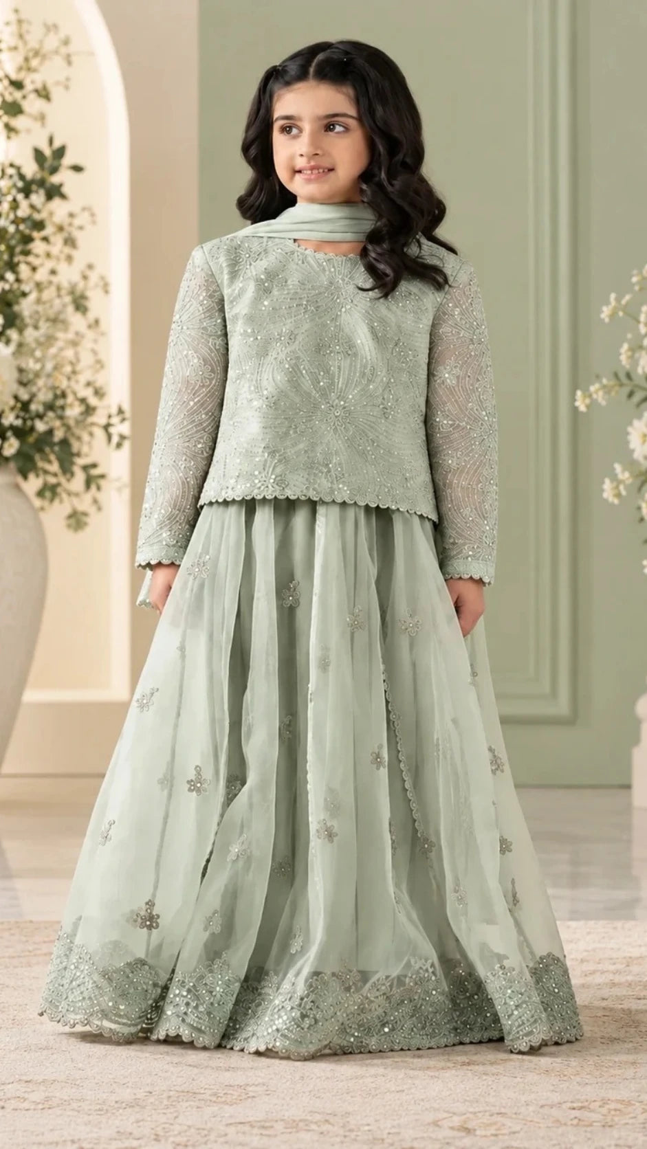 A young girl modeling a sage green traditional Indian outfit consisting of a long-sleeve embroidered jacket, a matching flared skirt with embroidered borders, and a coordinating dupatta, set against an elegant indoor backdrop.