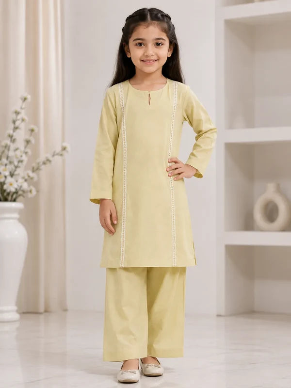 A young girl modeling a light yellow two-piece kurta and trouser set with white vertical embroidery, standing in a bright, modern indoor room.