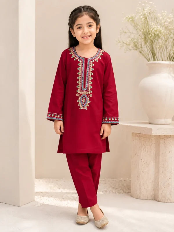 A young girl modeling a deep red kurta and pant set with intricate multicolor embroidery on the neckline and cuffs, standing in a warm, neutral-toned indoor setting.