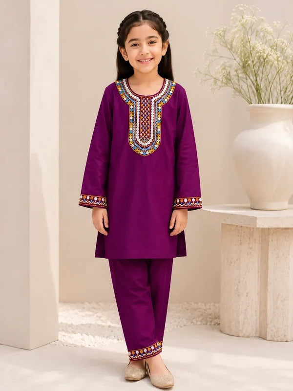 A young girl stands indoors wearing a purple traditional outfit with colorful embroidered detailing around the neckline and cuffs, paired with matching pants and beige shoes. A white vase with dried flowers sits on a stone side table in the background.
