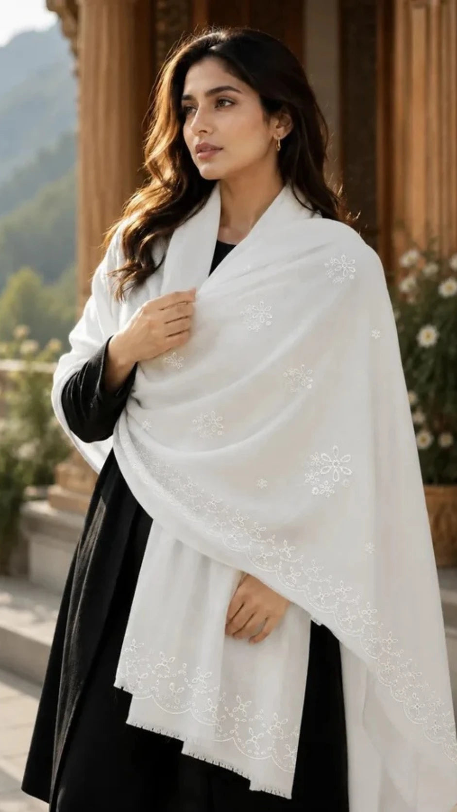A woman wearing a white embroidered shawl over a black dress, standing outdoors in front of a stone architectural background.