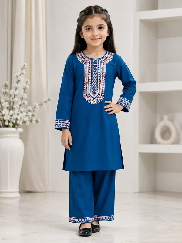A young girl modeling a royal blue two-piece kurta and pant set with colorful geometric embroidery around the neckline and cuffs, standing in a bright, modern room.