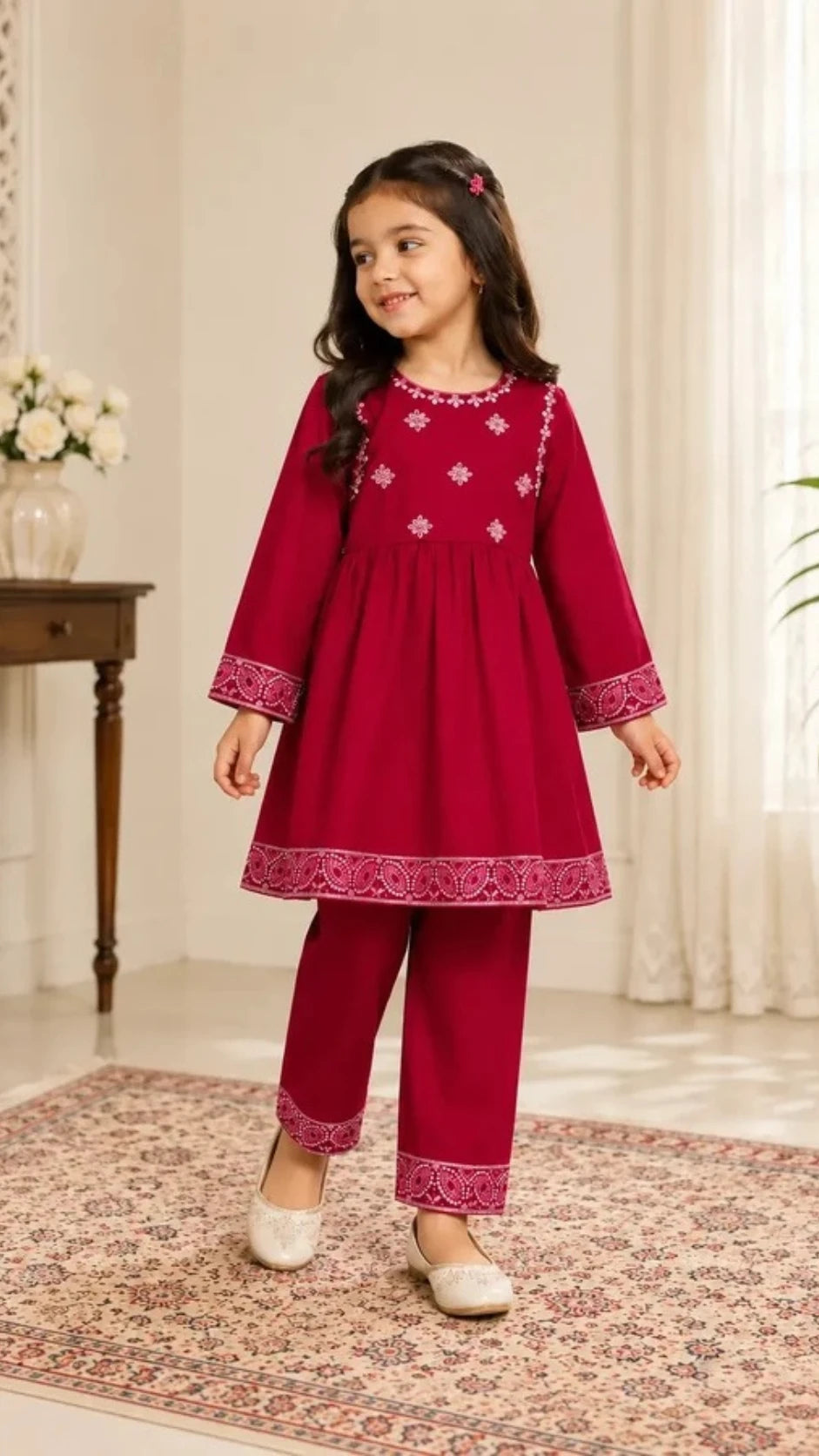 A young girl modeling a deep red traditional Indian outfit with white embroidery and matching red pants, standing on a patterned rug in an indoor setting.